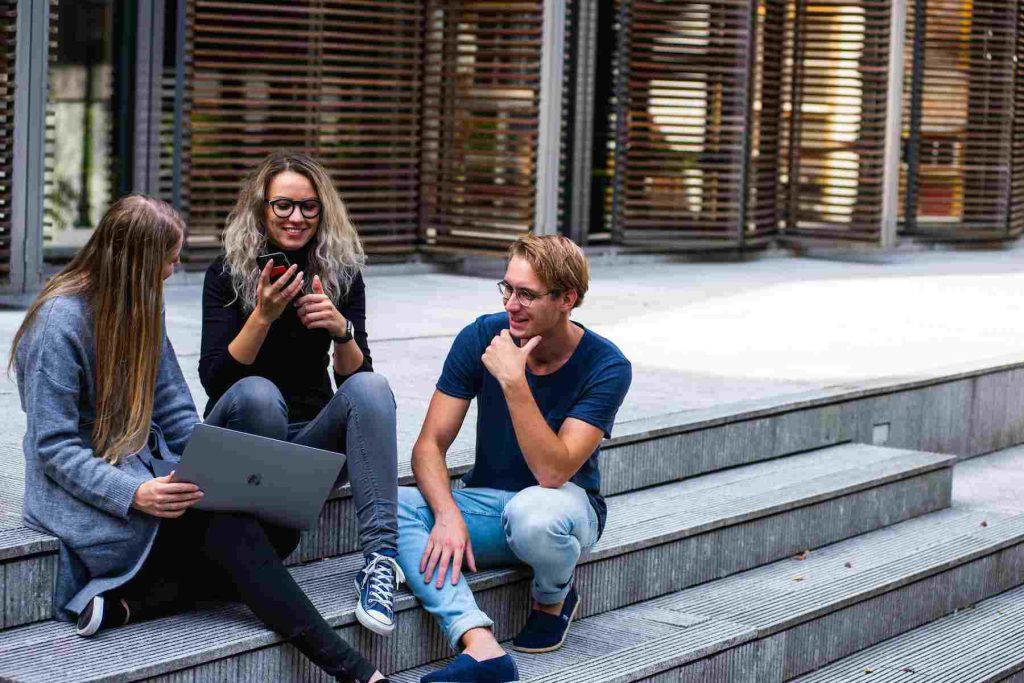 Three people smiling and talking while sitting on the stairs with laptop and cellphone