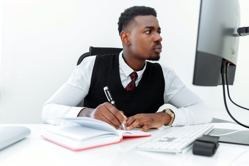 Man writing on his notebook while looking at his desktop monitor